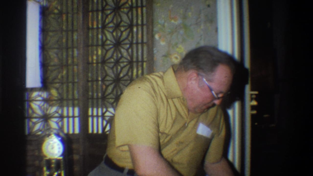 In a 1973 Troy, New York home-movie frame, an older man in a yellow shirt leans forward beside a chair in a dim room.
