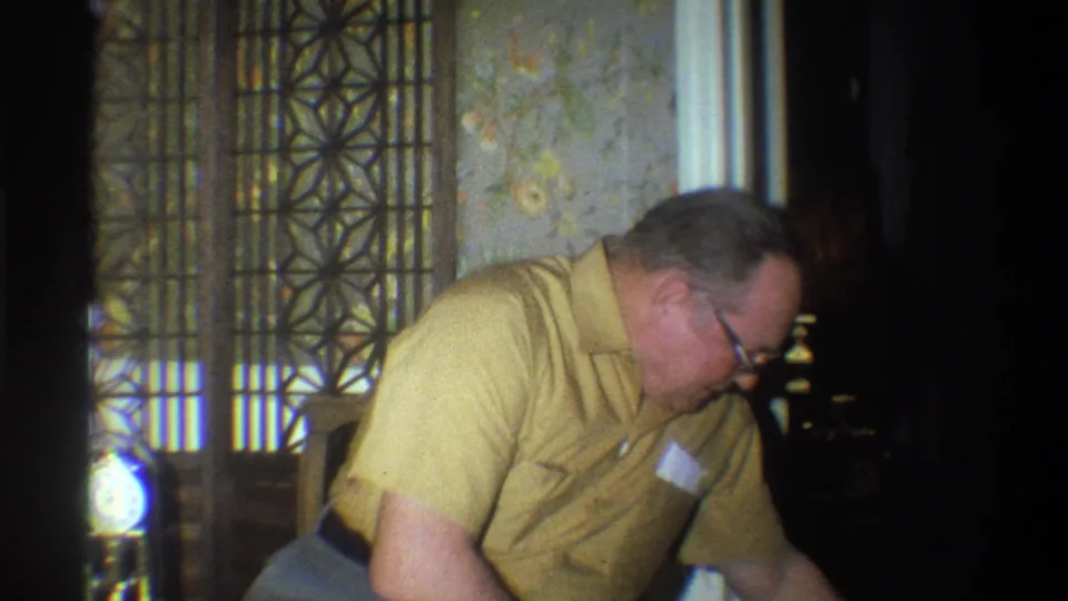 The man remains bent forward near a chair, with patterned wallpaper and a decorative screen visible behind him.