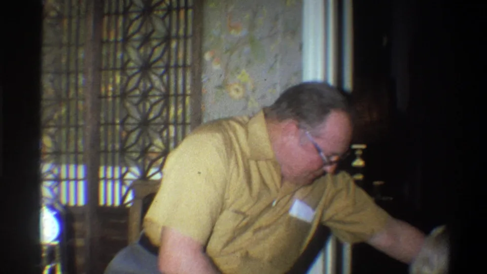 In the Troy home interior, the man reaches off-camera while seated or leaning beside a chair, suggesting informal interaction.
