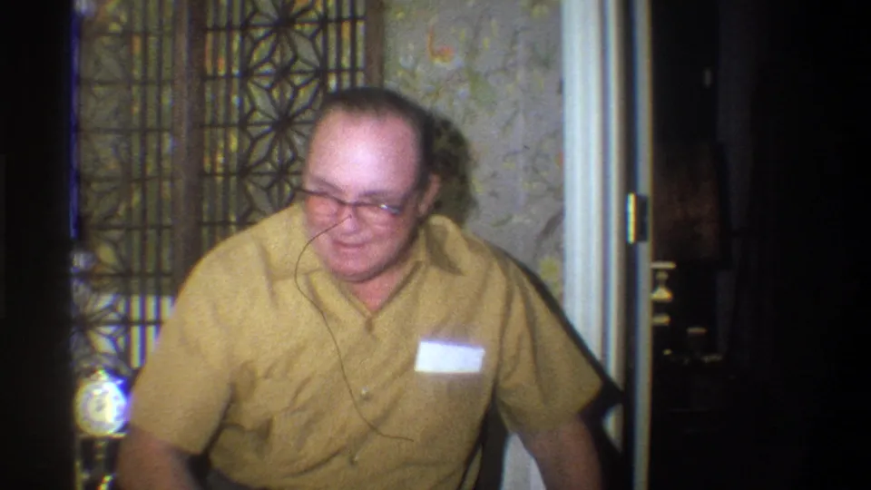 The man turns more toward the camera with a faint smile, framed by wallpaper, a decorative screen, and dark interior space.