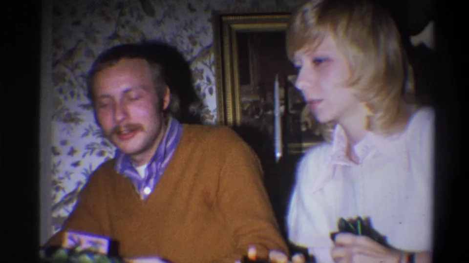 A mustached man in a brown sweater sits beside a blonde woman in a 1973 Troy, New York home interior.