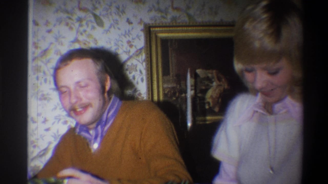 1973 Troy, New York home-movie frame showing a laughing seated man beside a blonde woman in a private interior with floral wallpaper.