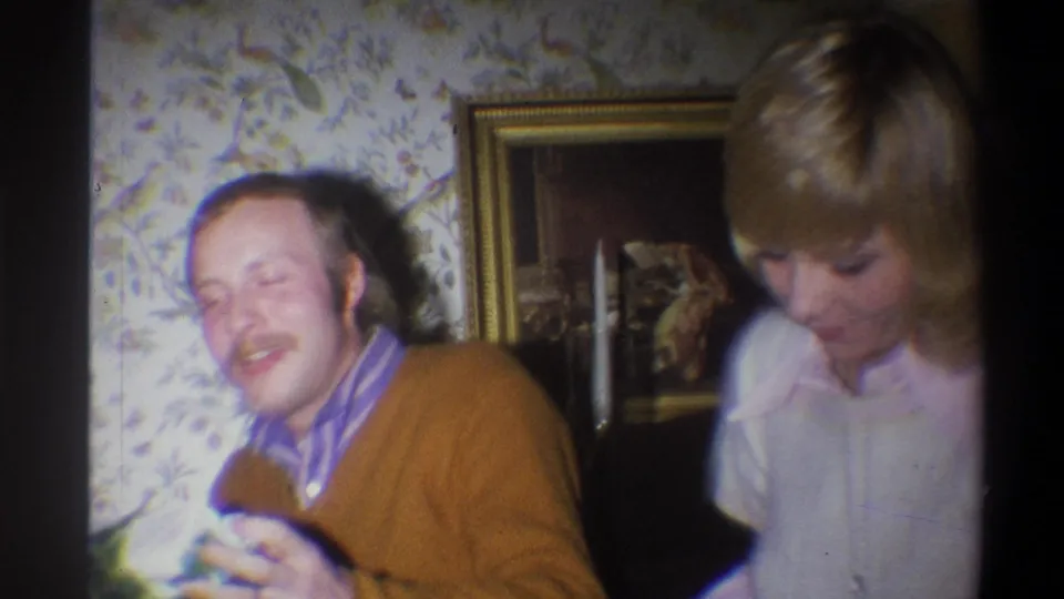 1973 Troy, New York 8mm frame of a seated man smiling near a blonde woman, with floral wallpaper and framed art behind them.