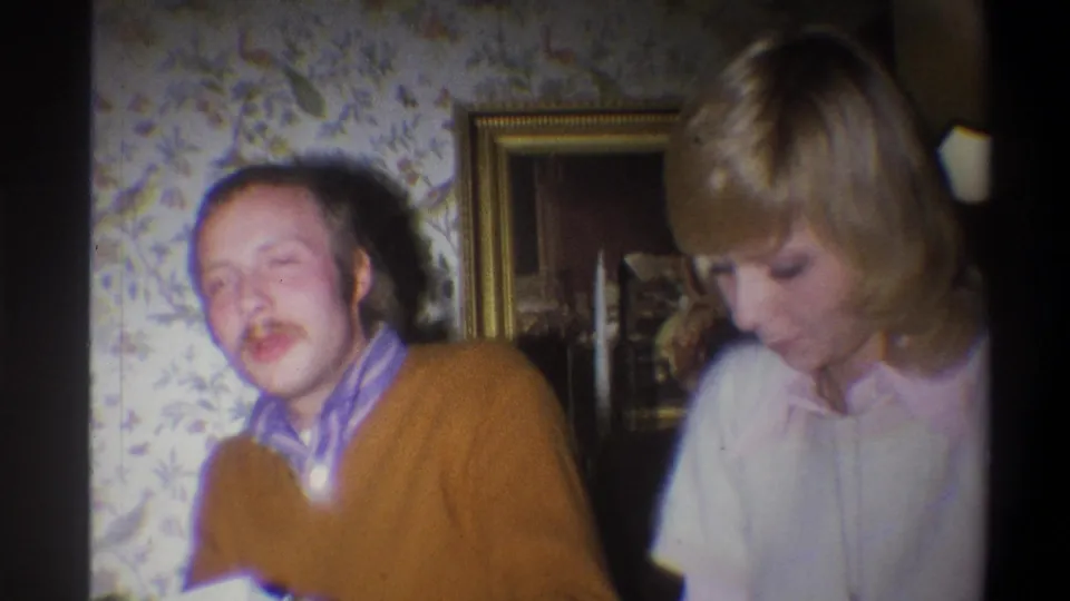1973 Troy, New York home-movie frame showing a man mid-expression beside a woman in a private room with patterned wallpaper.