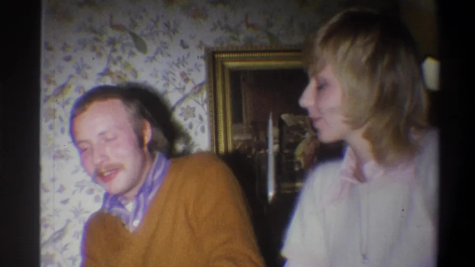 1973 Troy, New York 8mm frame showing a blonde woman turned toward a seated man in a cozy private interior.