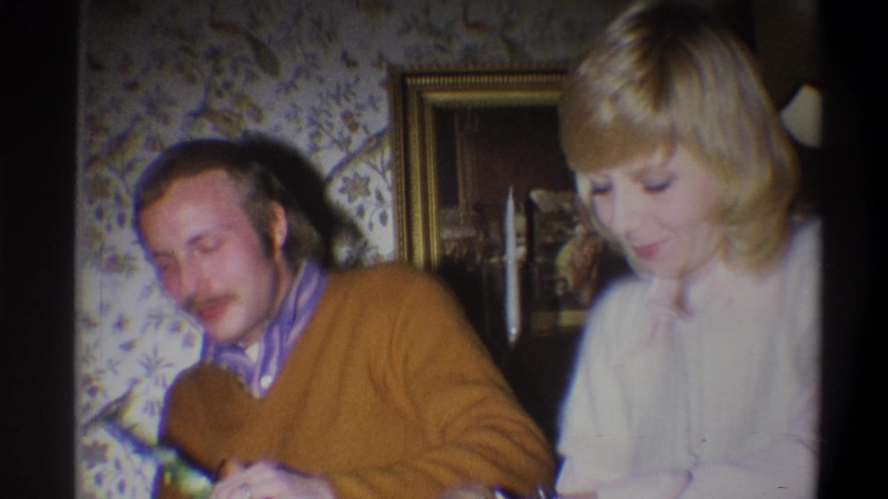 Man in a brown sweater sits beside a blonde woman in a softly lit room.