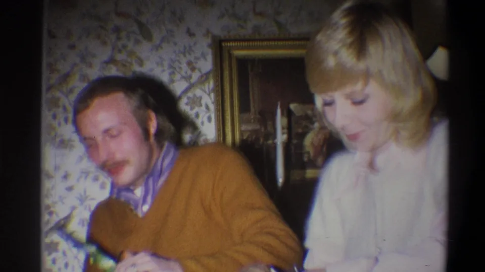 In a Troy, New York 1973 home-movie frame, a man in a brown sweater sits next to a woman in a pale blouse during a gift exchange.
