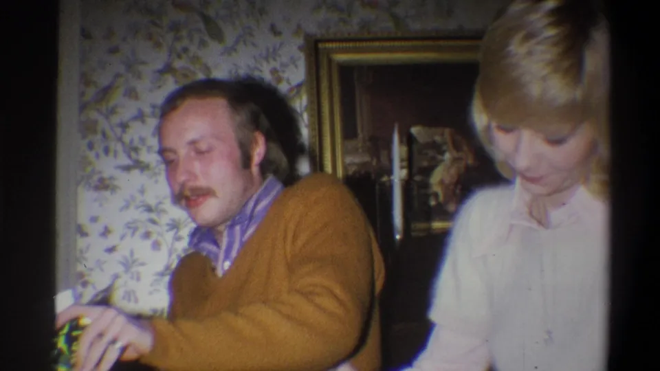 The man smiles during the 1973 home-movie gift exchange as the woman looks down beside him in the private interior.
