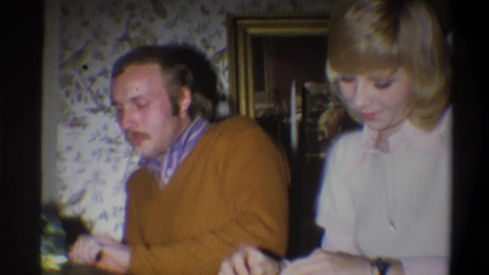 A 1973 Troy home-movie frame shows the couple seated close together while the woman focuses on the gift or wrapping in her hands.