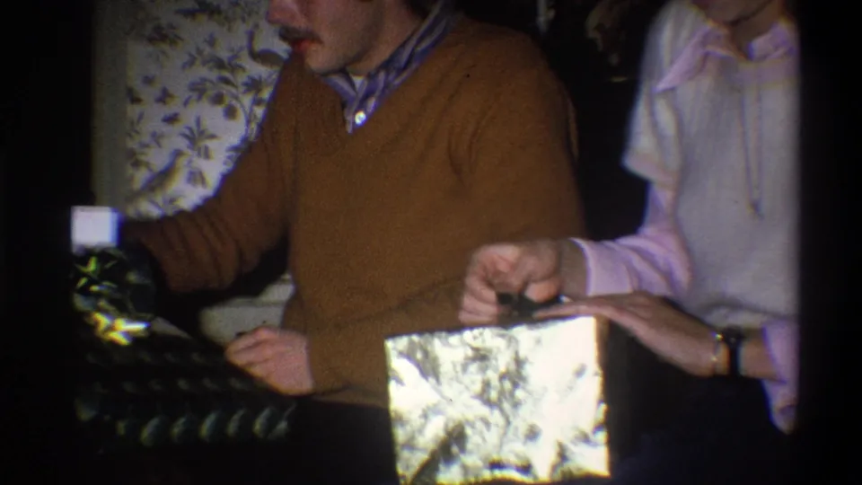 The woman holds reflective wrapping as the man handles a dark bottle or package during the Troy 1973 holiday exchange.