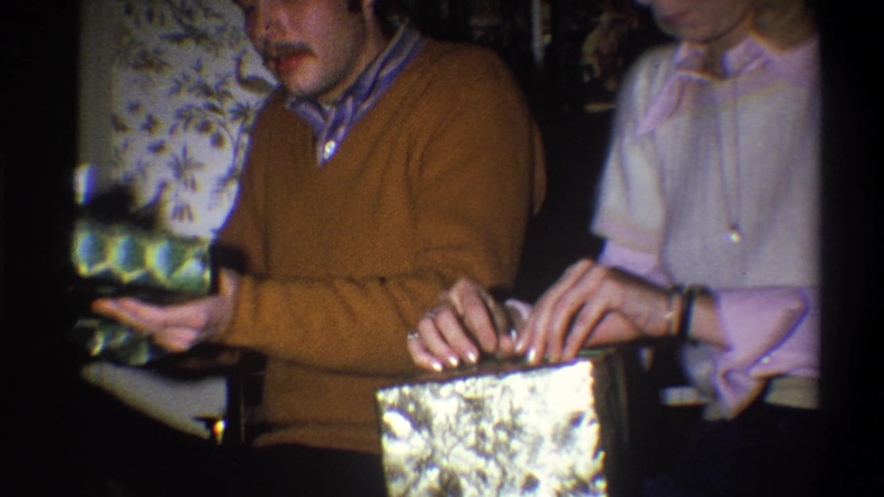In the supplied 1973 Troy, New York home-movie context, two adults sit by floral wallpaper while handling wrapped Christmas presents.