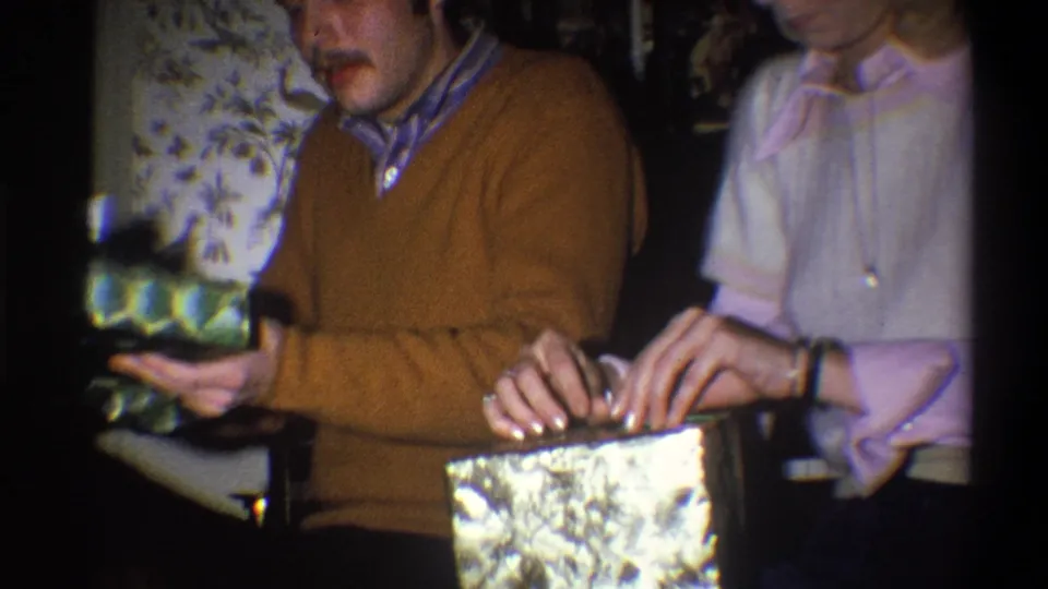 In the supplied 1973 Troy, New York home-movie context, two adults sit by floral wallpaper while handling wrapped Christmas presents.