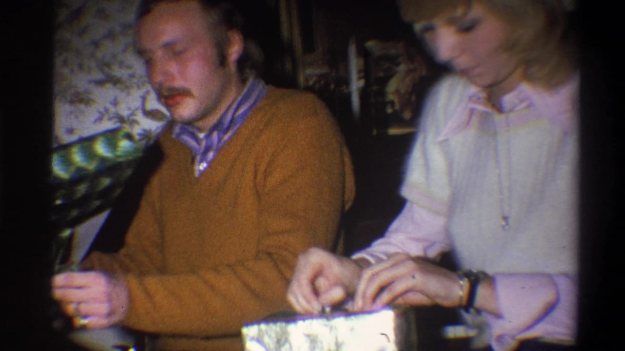 Woman unwraps a gift while a man in a brown sweater sits beside her in a dim interior.