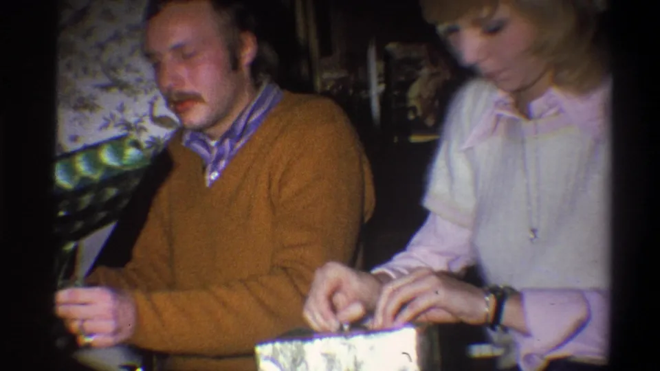 A woman in a pale blouse opens wrapping paper next to a man in a brown sweater during the documented 1973 holiday home movie.