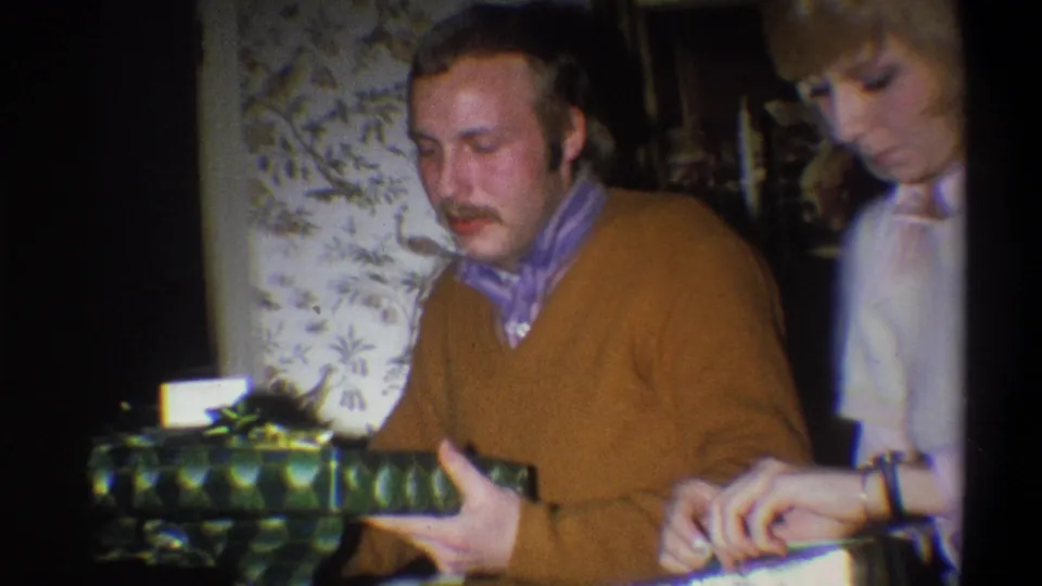 The man lifts or steadies a green wrapped present as the woman continues unwrapping, with patterned wallpaper behind them.
