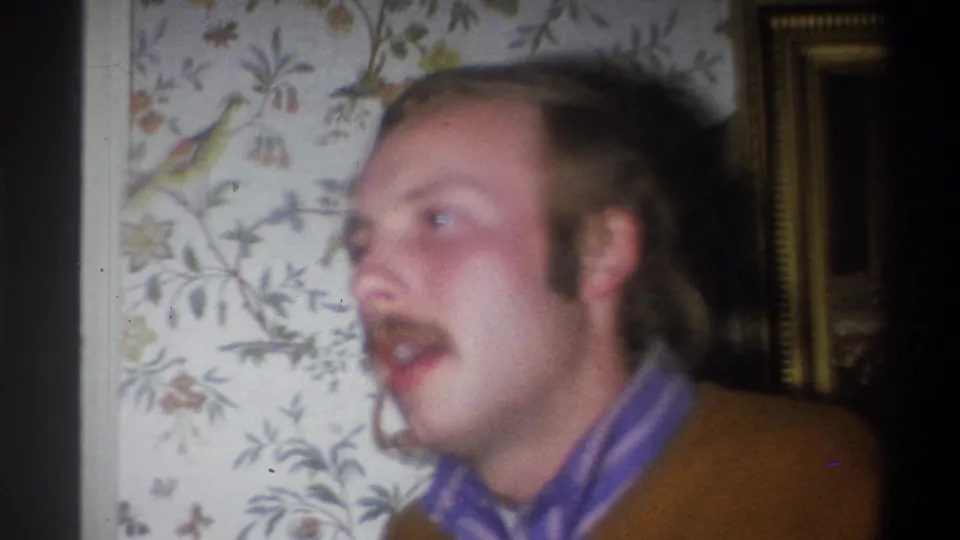 Close-up of the man in the brown sweater and purple shirt, seen against floral bird wallpaper in the 1973 home-movie setting.
