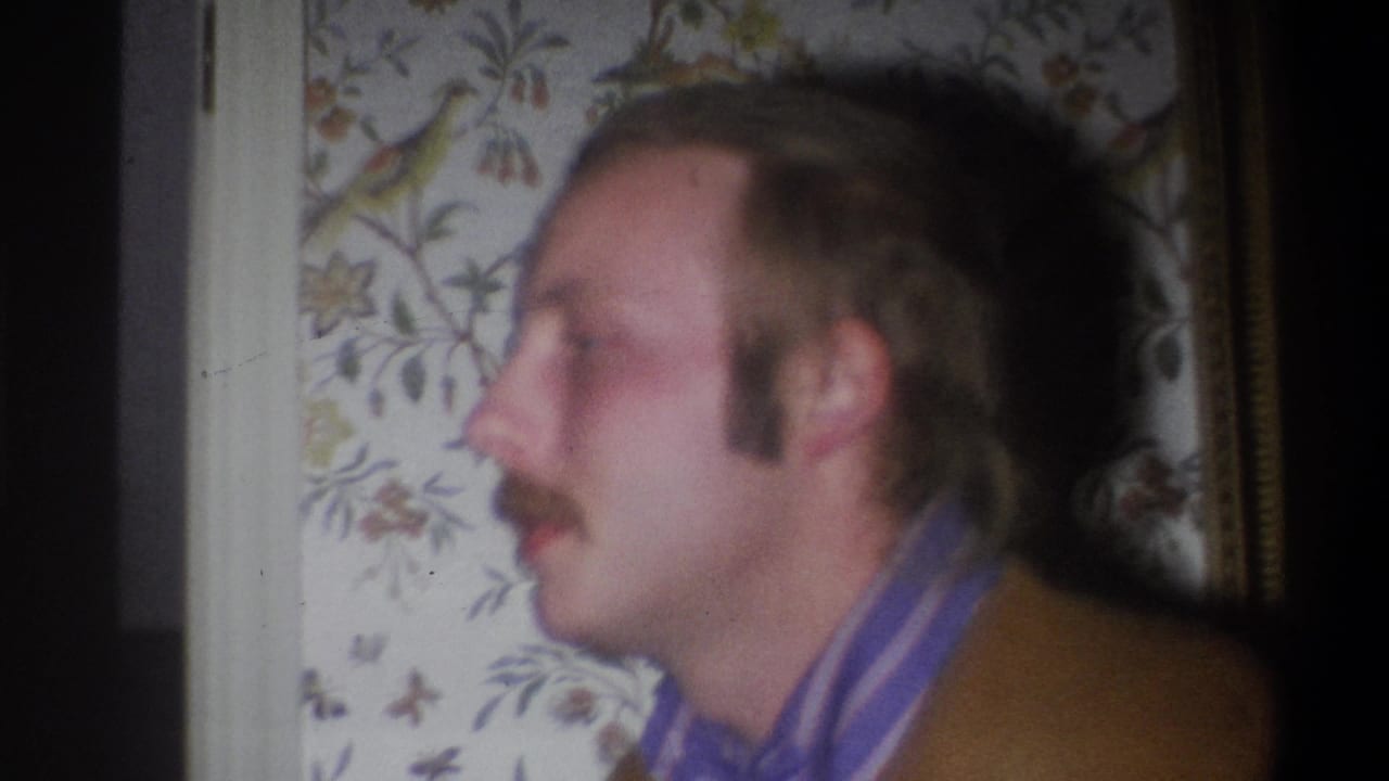 Mustached man in profile beside floral wallpaper and doorway trim.