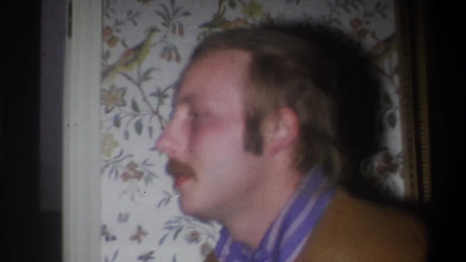 In a 1973 Troy, New York home-movie frame, a mustached man appears in profile near floral wallpaper and interior trim.