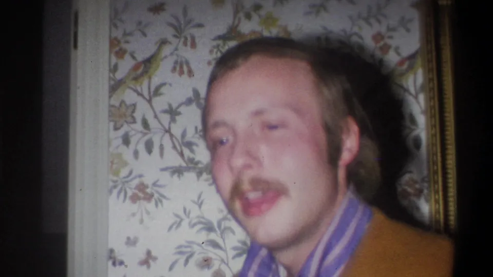 The man turns more toward the camera, wearing a brown jacket and striped shirt in a Troy, New York interior.