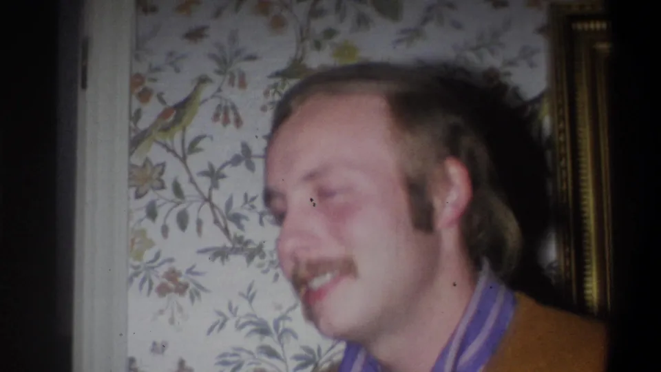 The man smiles in a close indoor 8mm frame, with floral wallpaper and wall trim behind him.