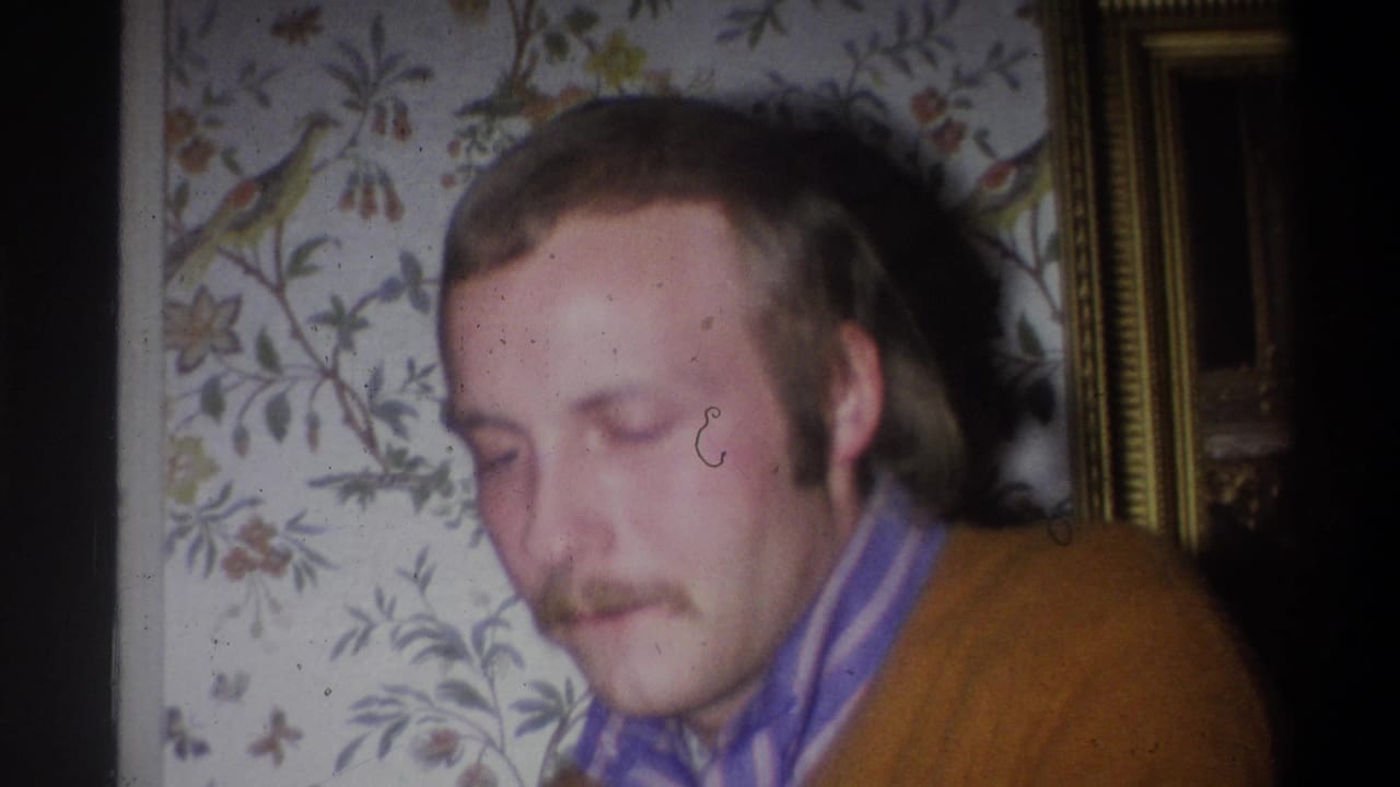 In a 1973 Troy, New York home-movie frame, a mustached man in a brown sweater and striped shirt looks downward beside floral wallpaper.