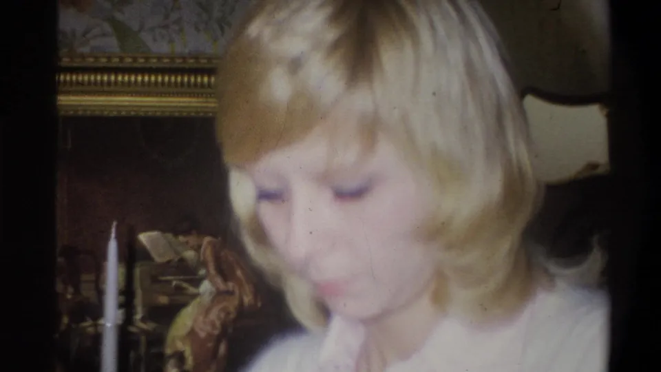 A blonde woman in a light top looks down in a 1973 Troy, New York home interior, with framed art and a lamp behind her.