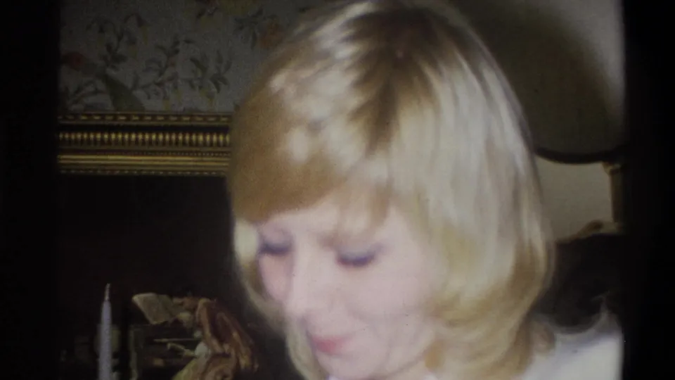 The blonde woman appears to smile faintly in a soft 1973 Troy, New York home-movie frame with framed art and patterned wallpaper visible.