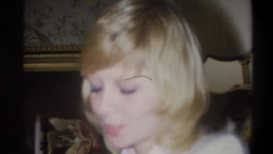 A blonde woman looks down with a slight smile in a 1973 Troy, New York private interior, with a lamp and framed picture behind her.
