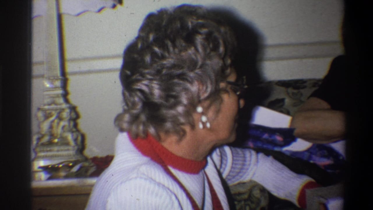 Older woman in glasses sits on a couch beside a lamp, turned toward someone off camera.