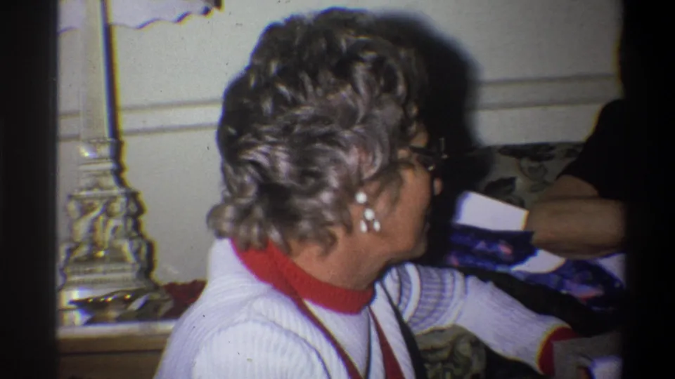 In a 1973 Troy, New York home-movie frame, an older woman in glasses, pearl earrings, a white sweater, and red turtleneck sits talking indoors.