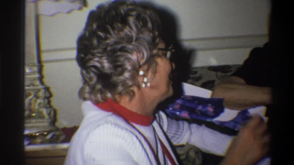 The woman remains seated in profile, appearing to speak or gesture, with another adult partly visible beside her in the private interior.