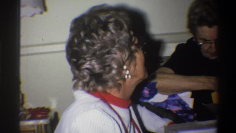 The home-movie frame shows the woman turned toward another adult with glasses, with the couch and lamp reinforcing the domestic interior setting.