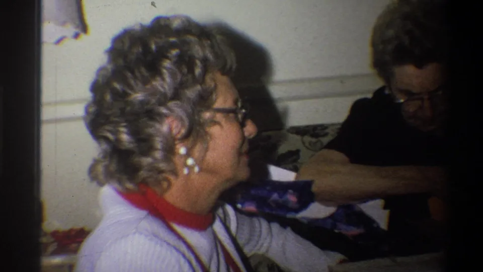 The woman smiles in profile while seated on the couch, with another adult nearby and soft 8mm grain visible across the indoor scene.