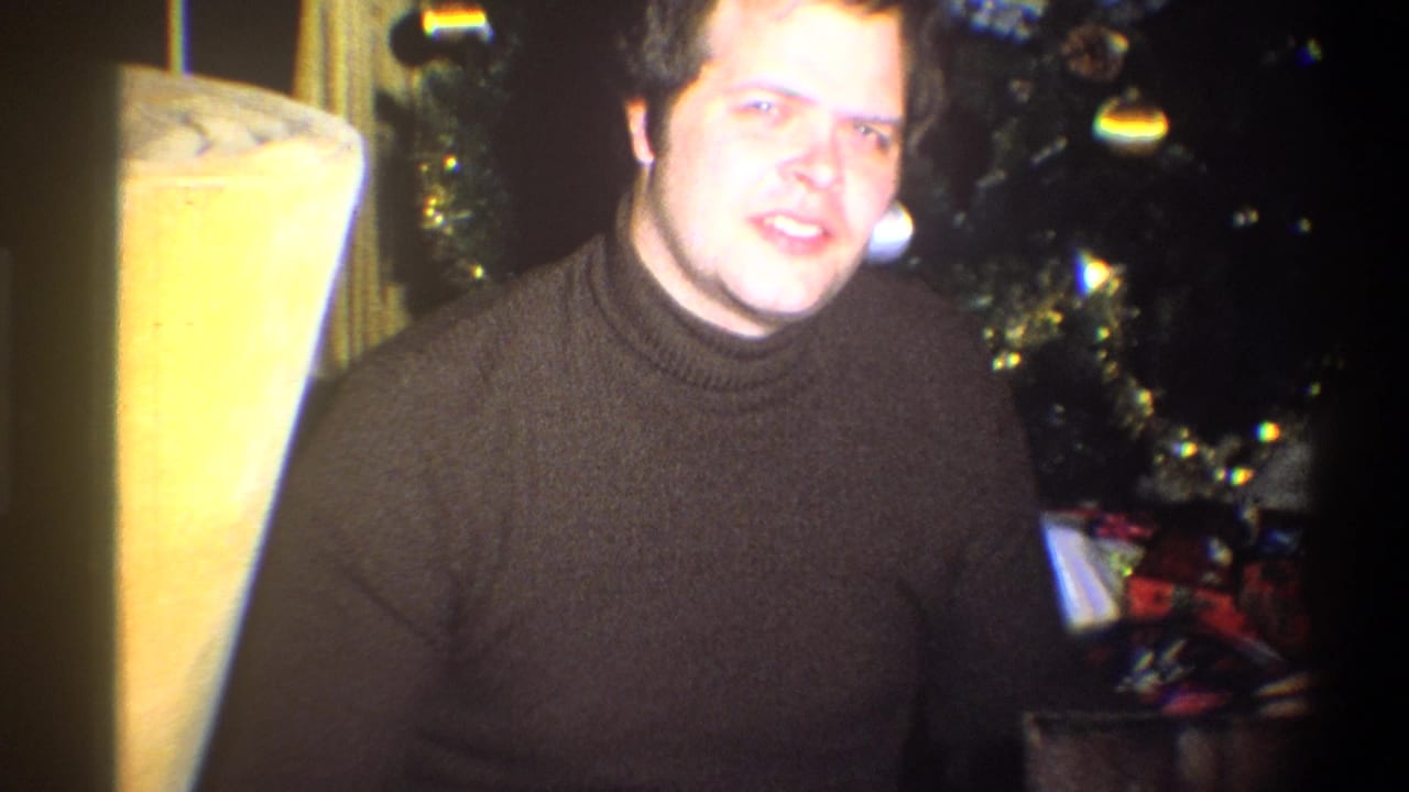 Man smiling toward the camera beside a Christmas tree and wrapped gifts.