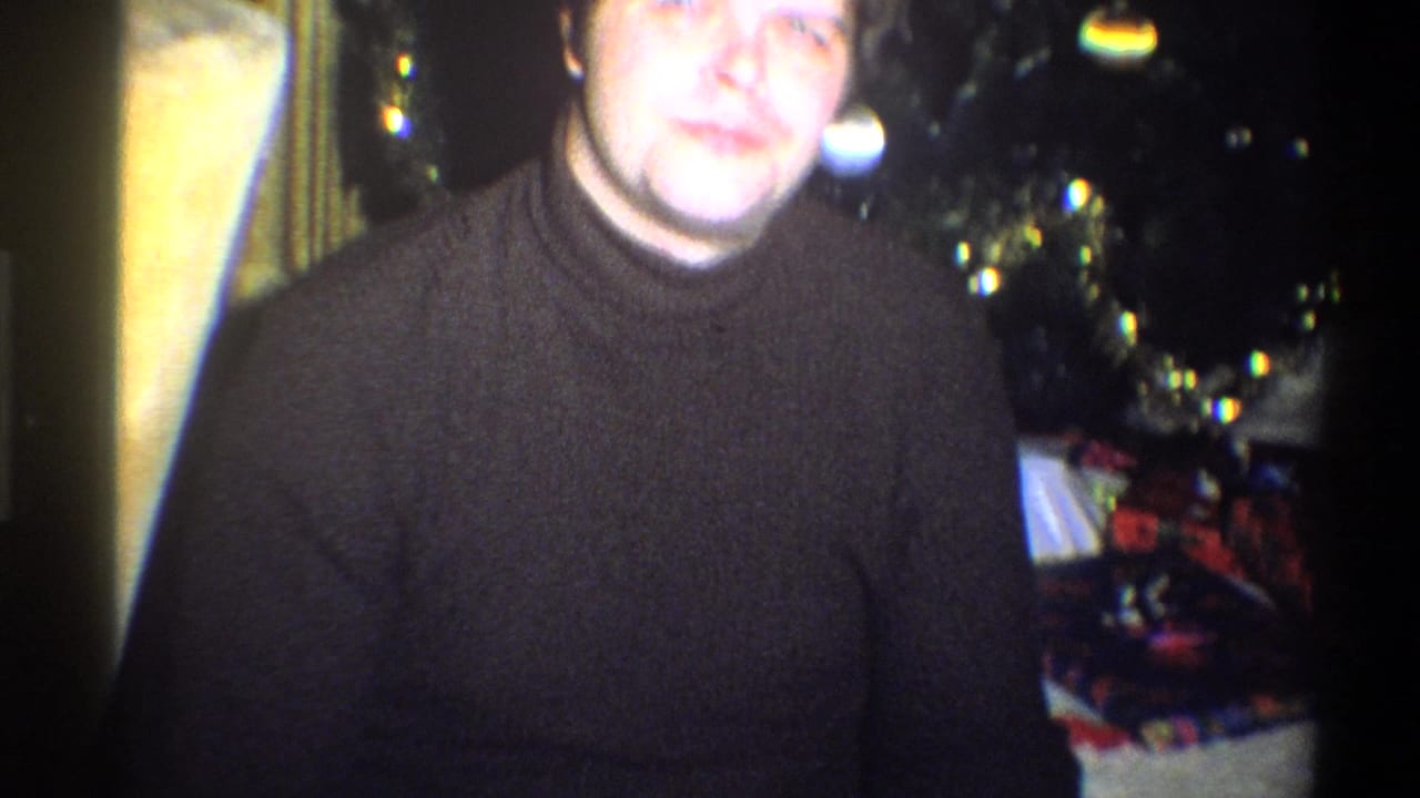 Man in a dark turtleneck seated beside a decorated Christmas tree with gifts behind him.
