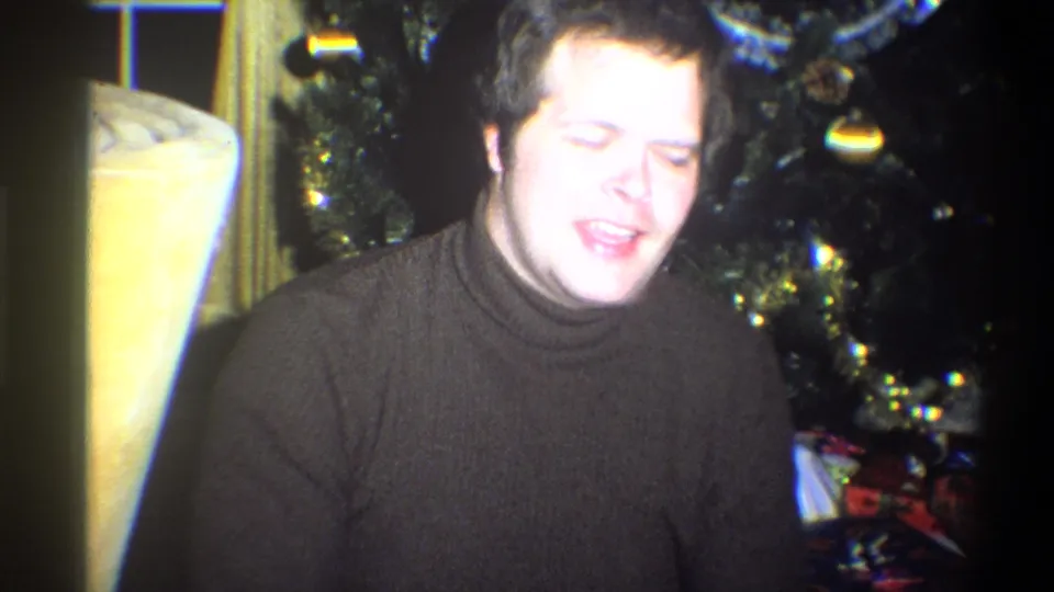 The man appears to smile or speak toward the camera near a Christmas tree in the Troy, New York holiday home interior.