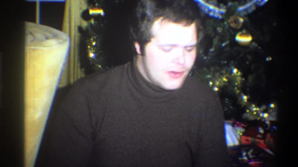 A 1973 home-movie frame shows the man looking down while seated beside holiday decorations and gifts in a private interior.