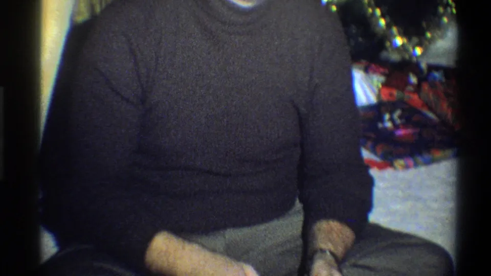 The camera frames more of the seated man's torso and lap, with the Christmas tree and wrapped gifts still visible behind him.