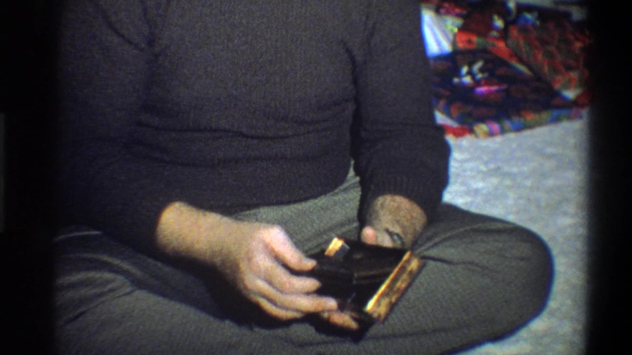 Man seated on carpet holding a small black box and wrapping near a Christmas tree.