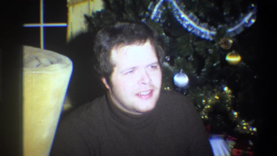 A man in a dark turtleneck sits near a decorated Christmas tree and upholstered chair in a 1973 Troy, New York home interior.