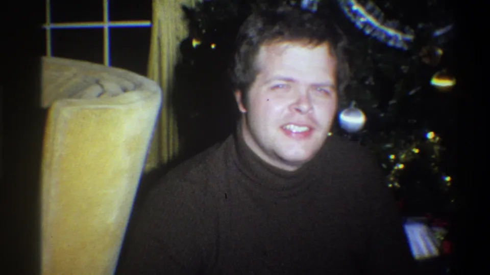 In 1973 Troy, New York, a smiling man sits in a living-room interior beside a Christmas tree with tinsel and ornaments.