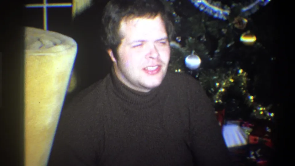 Young man in a dark sweater smiles near Christmas ornaments and a yellow chair in a private Troy, New York interior.