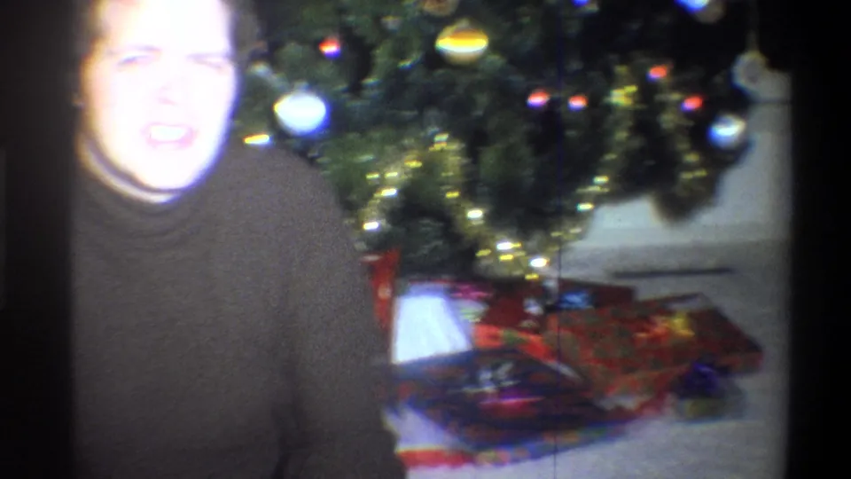 A bright, soft-focus frame shows the young man next to the Christmas tree with wrapped gifts below it.