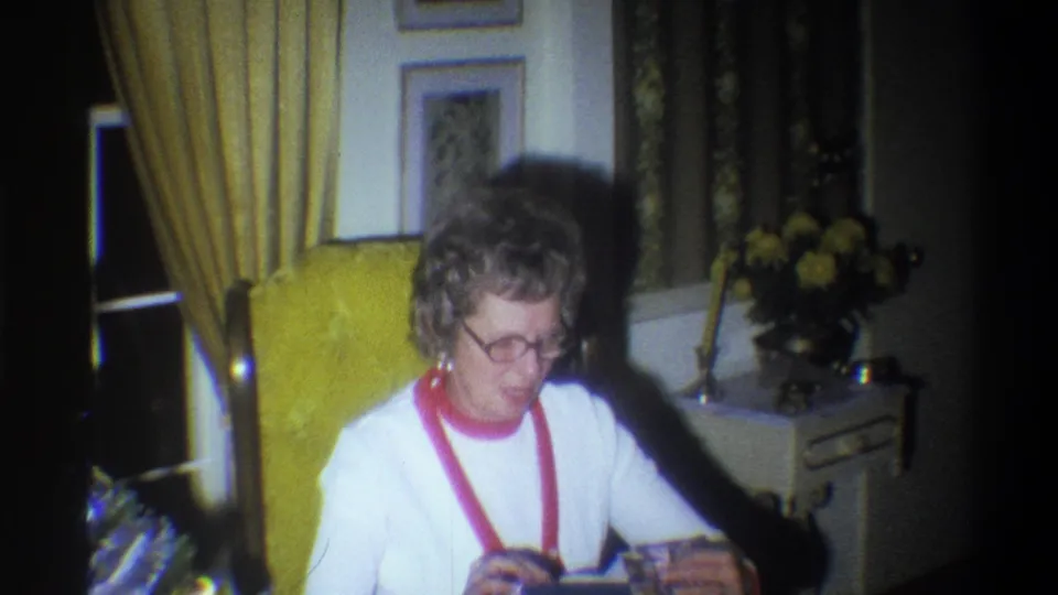 Older woman wearing glasses sits in a yellow armchair in a 1973 Troy, New York home interior, looking down at a small object.