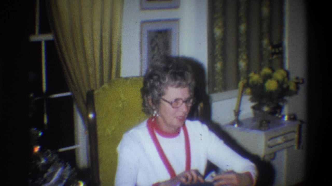 Elderly woman with glasses sits in a yellow armchair in a living room.