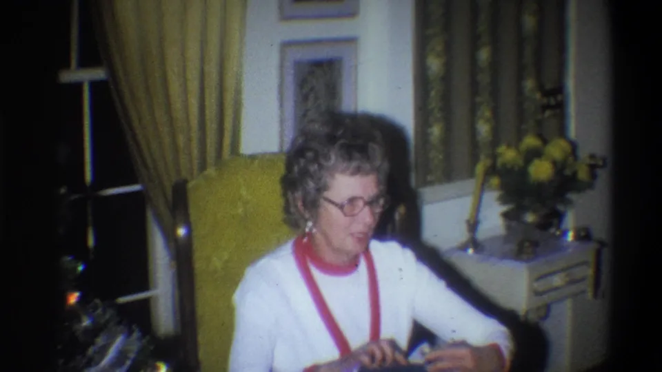In a 1973 Troy, New York home-movie frame, an elderly woman in glasses sits in a yellow armchair near curtains, framed pictures, and flowers.