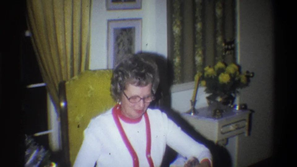 The seated woman looks down in a warmly lit 1973 Troy living room, with a yellow chair, drapes, framed pictures, and flowers behind her.