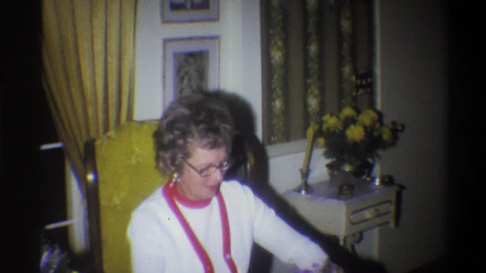 The woman leans forward in the yellow armchair, framed by curtains, wall art, and a side table with flowers in the private Troy interior.