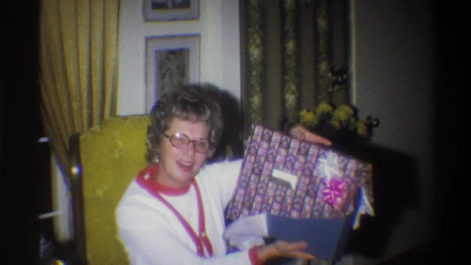 The elderly woman smiles toward the camera while holding a patterned Christmas gift box with a bow in the 1973 Troy home-movie scene.