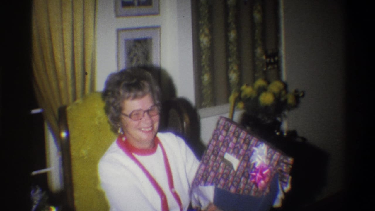 In a 1973 Troy, New York home movie frame, a woman in glasses smiles while holding a wrapped birthday gift in a warmly lit living room.